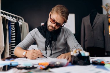 Moda gözlükleri takmış sakallı adam masaya çizimler çiziyor ve dikiş aletleri etrafa saçılıyor. Modern atölyede kıyafet tasarlayan bir terziydi..