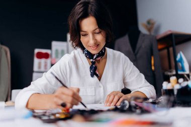 Yaratıcı, olgun terzi kendi dikiş atölyesinde çalışırken özenle çizimler yapıyor. Beyaz kadın şık giyinmiş, favori hobisinin tadını çıkarıyor..