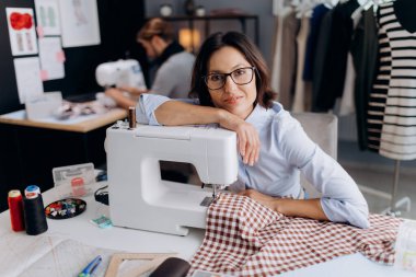 Gözlüklü, mavi gömlekli, iş yerinde dikiş makinesiyle oturan çekici, koyu renk saçlı kadının portresi. İnsanlar, terzilik ve yaratılış kavramı.