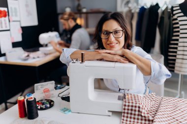 Gözlüklü çekici moda tasarımcısı başını dikiş makinesine dayamış ve içten bir şekilde gülümsüyor. Modern terzide çalışan, gözlüklü, beyaz, olgun bir kadın..