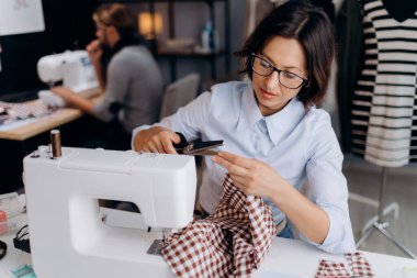 Gözlüklü deneyimli bir terzi modern atölyede oturuyor, kareli kumaşı terzilik makasıyla kesiyor ve dikiş makinesine dikiş dikiyor. Moda endüstrisi kavramı.