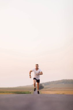 Spor elbiseli, spor ayakkabılı, dışarıda asfalt yolda koşan güçlü bir genç. Kırsal kesimde dayanıklılık için mesafe koşan yakışıklı beyaz adam..