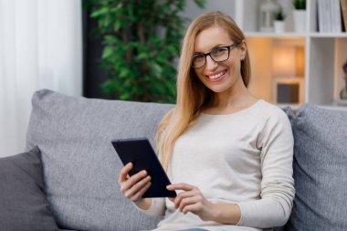 Elinde dijital tabletle kanepede oturan, göz giysisi içinde gülümseyen beyaz kadın portresi. İnsan kavramı, teknoloji ve modern yaşam tarzları.