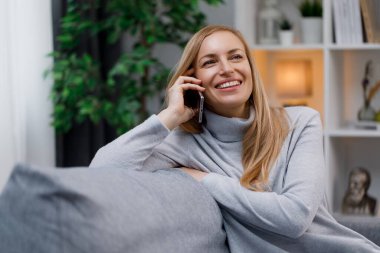Gündelik giysiler içinde gülümseyen beyaz kadın koltukta otururken modern akıllı telefondan hoş sohbetler ediyor. Mutlu olgun sarışın rahat oturma odasında konuşmak için cep telefonu kullanıyor..
