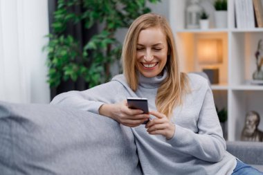 Rahat koltukta oturan, gündelik kıyafetler giymiş, modern akıllı telefonlu neşeli beyaz kadın. Boş zamanlarında sosyal ağları kaydıran mutlu olgun bir kadın..