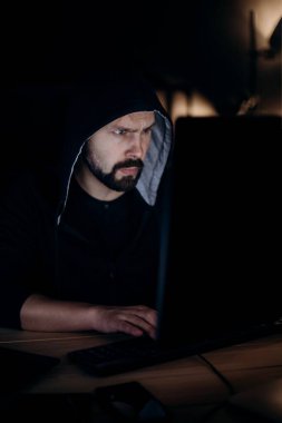 Gündelik kapüşonlu beyaz adam karanlık odada oturuyor ve modern bilgisayarda kodluyor. Kişisel bilgisayar kullanarak siber suç işleyen profesyonel bir hacker..