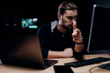 Siyah kapüşonlu profesyonel siber terörist etrafta bir sürü bilgisayarla karanlık odada otururken parmağıyla işaret ediyor. Hackleme ve teknoloji konsepti.