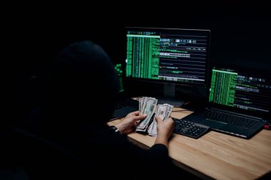 Kapüşonlu hacker 'ın karanlık odada birden fazla bilgisayarı var ve para sayıyor. Erkeklerin elindeki dolar banknotlarına odaklan.