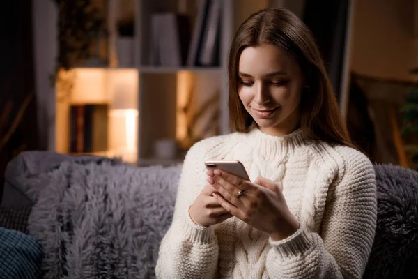 Uzun kahverengi saçlı, rahat koltukta oturan ve boş zamanlarında modern akıllı telefon kullanan beyaz bir kadın. İnsanlar, aletler ve ev yaşamları kavramı.