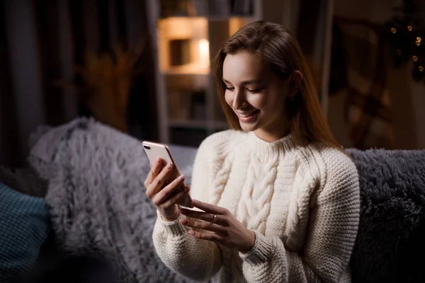 Örgü kazaklı sevimli beyaz kadın kanepede dinleniyor ve internette sörf yapmak için modern akıllı telefonu kullanıyor. Cihazlar ve yaşam tarzları kavramı.