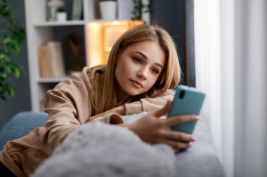 Sarı saçlı, rahat koltukta oturan ve seyyar ekrana bakan sıradan giyinmiş genç bir kadın. İnsan kavramı, teknoloji ve ev yaşamları.