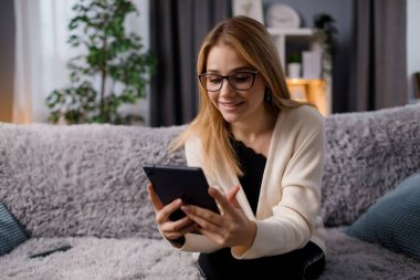 Kanepede oturan ve tablet ekranına bakarken gülümseyen sevimli genç kadın. Sıradan kıyafetler ve gözlük takan beyaz kadın sarışın..