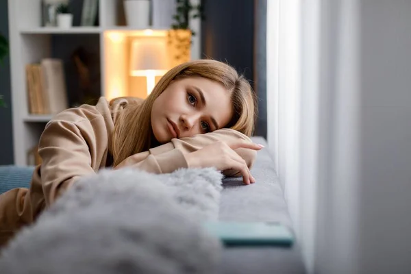 Stock image Attractive young woman in casual attire leaning with head on soft grey couch while resting during free time. Daydreaming and relaxation concept.