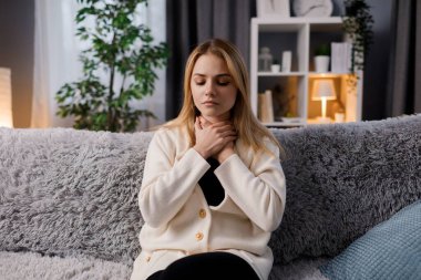 Mutsuz genç bir kadın kendini kötü hissediyor ve oturma odasında kanepede otururken boğaz ağrısı çekiyor. İnsanlar, soğuk ve sağlık sorunları kavramı.