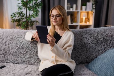 Rahat bir oturma odasında kanepede otururken dijital tablet kullanan gözlüklü hoş bir kadın. Modern teknolojiye sahip ev yaşamları.