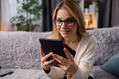 Elinde dijital tabletle rahat koltukta dinlenen sarı saçlı çekici genç bir kadının portresi. Gözlüklü güzel beyaz kadın evdeki modern cihazda internette sörf yapıyor..