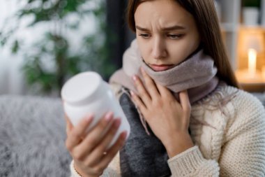 Evdeki kanepede otururken, sarı saçlı, ilaç kutusuyla muayene eden hoşnutsuz genç bir kadın. Sıcak eşarba sarılı hasta bir kadın boğaz ağrısı sırasında ilaç alıyor..