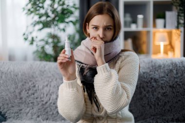 Sıcak bir eşarpla kanepede oturan ve elinde burun spreyi tutan üzgün beyaz kadın portresi. Mevsimsel grip sırasında burnu akan hasta genç bir sarışın..