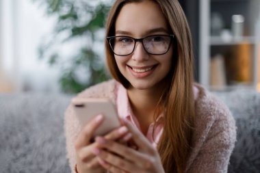 Pozitif beyaz kadın portresi rahat bir kanepede oturuyor ve kişisel akıllı telefonu elinde tutuyor. Gözlüklü genç bayan modern aletlerle internette sörf yapıyor..