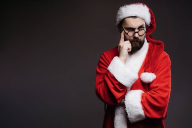 Kırmızı noel kostümlü düşünceli olgun bir adam stüdyoda karanlık geçmişiyle poz verirken parmağını şakağın yakınında tutuyor. Noel Baba şüphe içinde.
