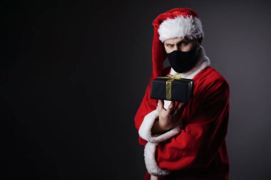 Kırmızı Noel Baba kostümlü ve siyah maskeli kendine güvenen olgun bir adam stüdyoda hediye kutusu tutuyor. Kış tatillerinde salgınla hediyeler.