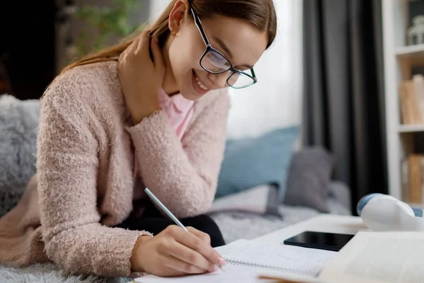 在家里读书的时候 戴着眼镜笑一个高加索女人 在笔记本上随意写字 勤勉的女学生利用业余时间准备考试 — 图库照片