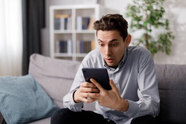 Kanepede otururken tablet ekranda şaşırmış yüz ifadesi olan beyaz bir adam. Sıradan giyinmiş bir adam evde beklenmedik haberler alıyor..