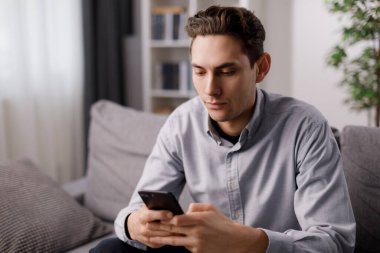 Kanepede otururken modern akıllı telefonlarda mesajlar yazan sıradan giyinmiş genç bir adam. Koyu saçlı beyaz adam evde otururken internette sohbet ediyor..