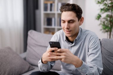 Rahat bir koltukta otururken modern akıllı telefon kullanan yüzü gülen yakışıklı beyaz bir adam. Rahat bir dairede boş zamanlarında online sohbetten hoşlanan genç adam..