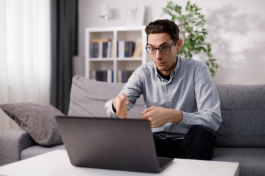 Gözlüklü ve günlük kıyafetleri olan genç bir adam kanepede oturuyor ve taşınabilir laptopta görüntülü sohbet yapıyor. Serbest çalışanların çevrimiçi toplantısı.