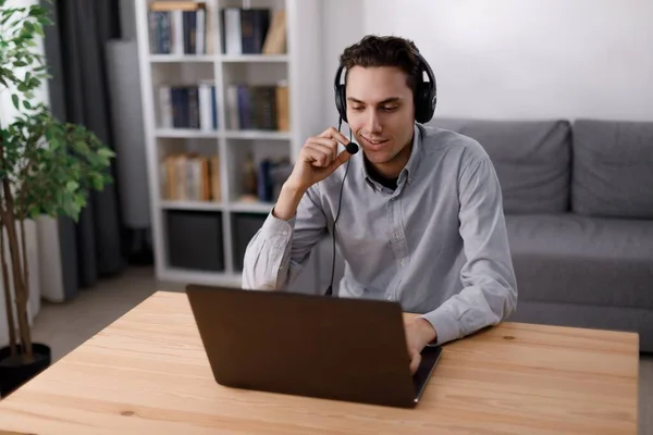 Evde video konferansı için modern kulaklık ve dizüstü bilgisayar kullanan beyaz bir erkek. Gündelik giyinen genç adam uzaktan çalışma sırasında online toplantı yapıyor..