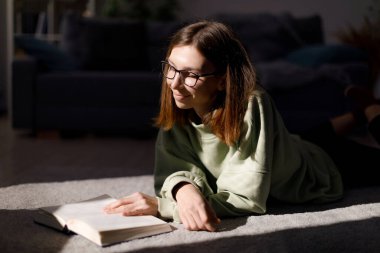 Çekici bir kız öğrenci yerde yatıyor ve açık bir kitaba bakıyor. Kafkasyalı genç bir kadın kendini eğitmek için rahat oturma odasında boş vakit geçiriyor..