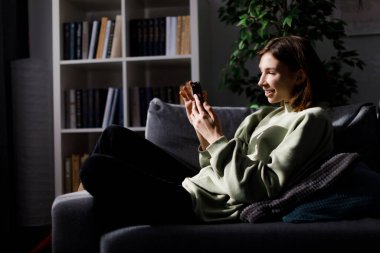 Evde rahat bir kanepede dinlenirken kişisel akıllı telefon kullanan rahat kapüşonlu genç esmerin yan görüntüsü. Sosyal ağ, gençlik ve modern aygıtlar kavramı.