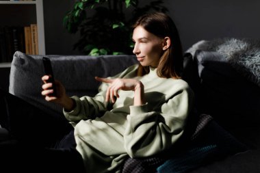 Rahat koltukta rahat rahat uyuyan ve video görüşmesinde parmağıyla ekranı gösteren alımlı genç bir kadın. Çevrimiçi iletişim, insanlar ve teknoloji kavramı.