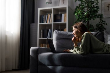 Rahat oturma odasındaki kanepede dinlenen gülümseyen genç bayan ve interneti taramak için modern dizüstü bilgisayar kullanıyor. Kafkasyalı kadın gündelik kıyafetlerle evde modern aletlerle vakit geçiriyor..