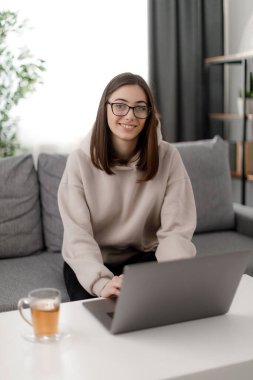 Gözlüklü, gülümseyen genç bir kadının oturma odasında dizüstü bilgisayarda ders izlediği bir manzara. Kahve masasının yanındaki kanepede bilgisayar ve bir fincan çayla oturan tatlı esmer kız..