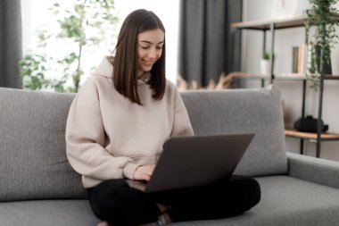 Lotus pozisyonunda kanepede otururken taşınabilir laptop kullanan mutlu beyaz öğrenci. Konsantre kadın daktilo ediyor ve dikkatlice ekrana bakıyor. Eğitim kavramı.