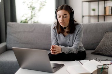 Modern dizüstü bilgisayar ve kulaklıkla rahat bir koltukta otururken video dersi almaya odaklanmış genç bir kadın. Günlük giysiler içinde beyaz bir kız öğrenci evde eğitim için zaman harcıyor..