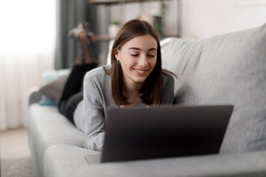 Rahat giyimli, çay içen ve kanepede dinlenirken film izleyen pozitif bir genç kadın. Evde boş zamanlarında modern laptop kullanan güzel beyaz tenli esmer..