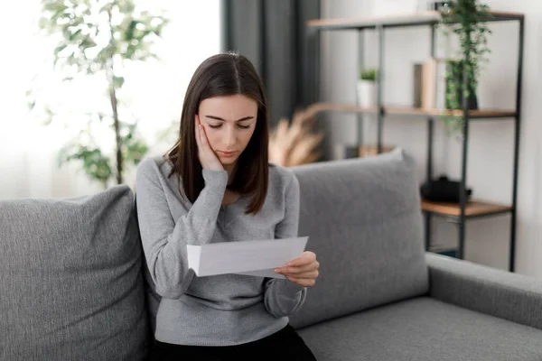 Serious brunette student supporting cheek with one hand while thoughtfully looking on document. Worried female in casual clothing sitting on sofa in living room while reading letter.