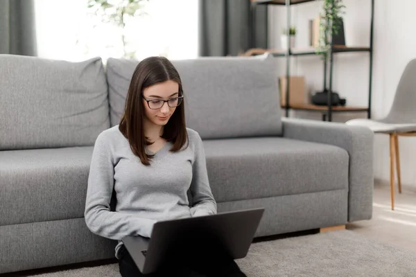Caucasian student writing essay on computer while sitting on floor in living room. Focused female wearing eyeglasses using wireless laptop for studying online.