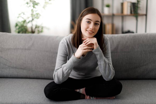 Pretty caucasian woman in casual attire sitting on sofa in lotus pose and leaning with hands on knees. Dark haired lady relaxing in living room during free time.