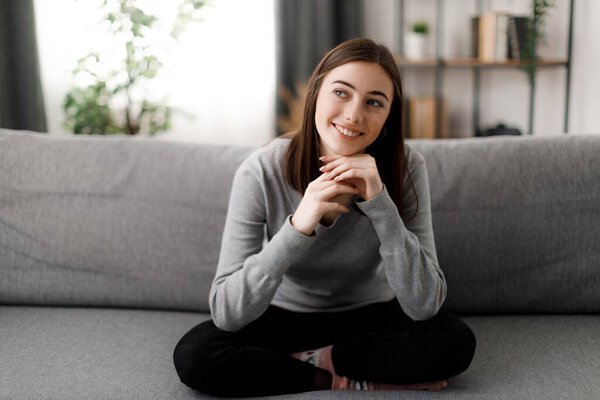 Dreamy young woman with brown hair sitting on comfy couch and looking aside. Caucasian young female in casual clothes enjoying leisure time at home.