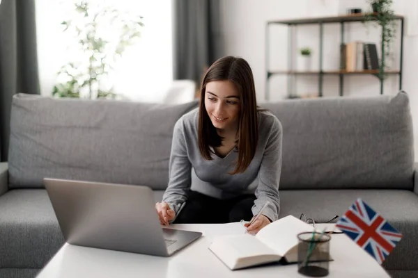 stock image Focused woman with happy smile on face studying on laptop in living room. Caucasian young female writing down in notebook during online english lesson.