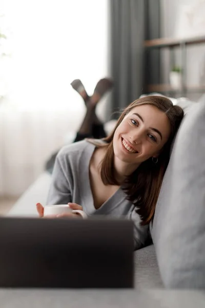 Smiling young woman in casual wear resting on comfy couch with hot drink and wireless laptop. Domestic lifestyles with modern gadgets.