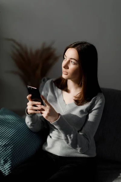 stock image Caucasian young woman thoughtfully looking aside while holding modern smartphone in hands. Dark haired female sitting on sofa and surfing internet on cell phone.