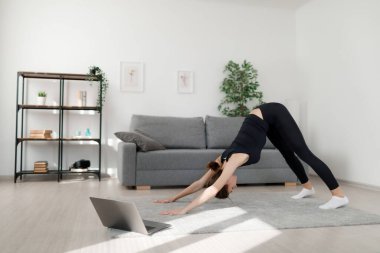 Spor elbiseli, aktif beyaz kadın modern dizüstü bilgisayar kullanırken halı üzerinde esneme egzersizleri yapıyor. Teknoloji ve iç eğitim kavramı.