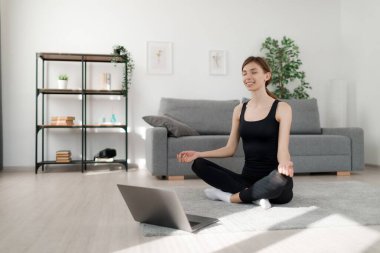 Siyah spor kıyafetleri içinde huzurlu genç bir kadın ev içi antrenman sırasında nilüfer pozisyonunda gözleri kapalı meditasyon yapıyor. İnternet 'te fitness videoları izlemek için kablosuz laptop kullanan Kafkas esmer.