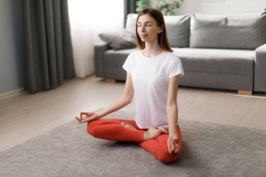 Huzurlu genç kadın yerde gözleri kapalı otururken yoga yapıyor. Kafkasyalı esmer, sabah evde çalışırken lotus pozisyonunda meditasyon yapıyor..