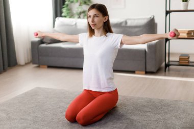 Beyaz tişörtlü ve kırmızı taytlı beyaz bir kadın aile içi eğitim sırasında dambıl kaldırıyor. Koyu renk saçlı, formda bir kadın yerde oturuyor ve antrenmanların keyfini çıkarıyor..